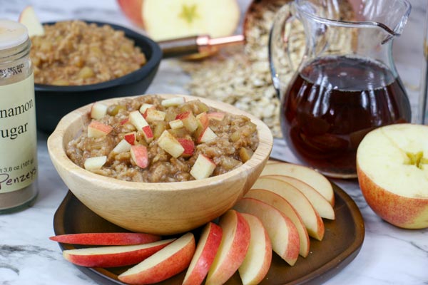apple cinnamon oatmeal in bowl on plate with apple slices. maple syrup, oats, apple halves, and cinnamon sugar topping surrounding