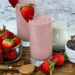 two glasses filled with frozen strawberry smoothie with fresh strawberries, almond milk, dates, and almond butter around