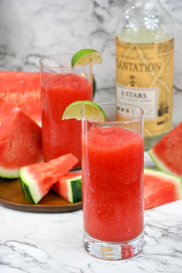 two glasses of boozy watermelon slush with watermelon wedges and a bottle of white rum in the background